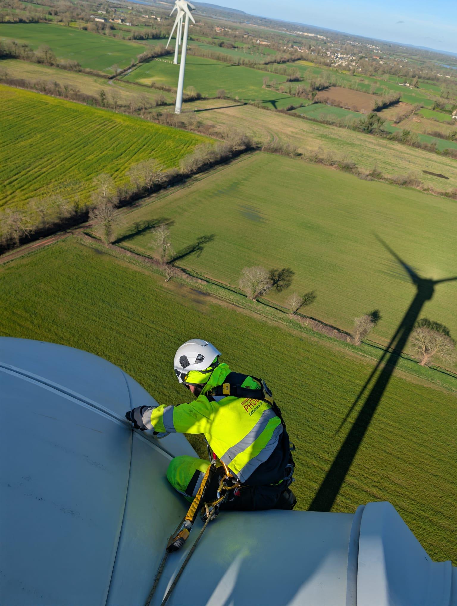 Phoenix staff in safety gear inspecting the top of a wind turbine above open green fields.